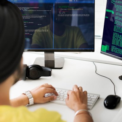 Person Schreibtisch Eine Person sitzt am Schreibtisch und tippt am PC, auf den Bildschirmen sieht man Code.