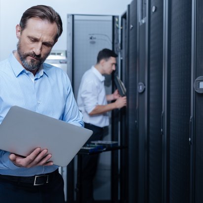 Zwei Männer im Rechenzentrum Zwei Männer stehen in einem Rechenzentrum, einer tippt etwas in den Laptop, den er hält.